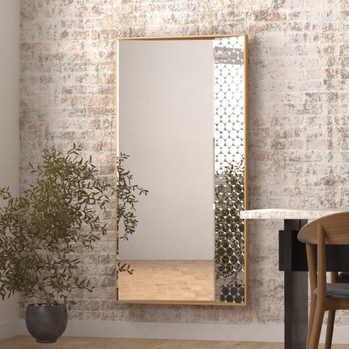 Modern rectangular mirror with a slim gold frame hanging on a light-colored, textured brick wall in a cozy room. A green leafy potted plant sits beside the mirror, and part of a wooden chair and dining table is visible, adding a warm, minimalistic touch to the space. Ideal for enhancing natural light and creating a sense of openness in the room