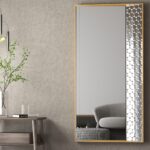 Contemporary rectangular mirror with a sleek gold frame and patterned edge, mounted on a textured gray wall. A minimalist wooden side table with a glass vase holding green branches and a book sits next to the mirror. Overhead, a modern pendant light adds a sophisticated ambiance. A reflection of a gray couch and checkered ottoman enhances the room’s stylish, cozy decor