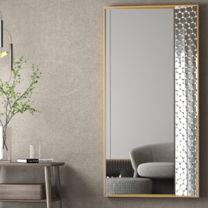 Contemporary rectangular mirror with a sleek gold frame and patterned edge, mounted on a textured gray wall. A minimalist wooden side table with a glass vase holding green branches and a book sits next to the mirror. Overhead, a modern pendant light adds a sophisticated ambiance. A reflection of a gray couch and checkered ottoman enhances the room’s stylish, cozy decor