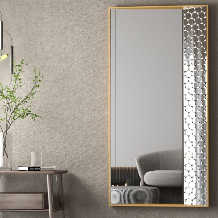 Contemporary rectangular mirror with a sleek gold frame and patterned edge, mounted on a textured gray wall. A minimalist wooden side table with a glass vase holding green branches and a book sits next to the mirror. Overhead, a modern pendant light adds a sophisticated ambiance. A reflection of a gray couch and checkered ottoman enhances the room’s stylish, cozy decor