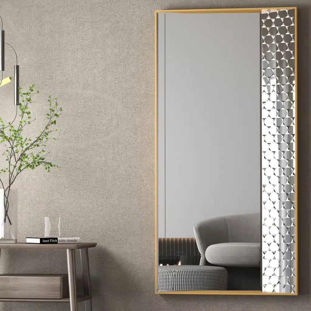 Contemporary rectangular mirror with a sleek gold frame and patterned edge, mounted on a textured gray wall. A minimalist wooden side table with a glass vase holding green branches and a book sits next to the mirror. Overhead, a modern pendant light adds a sophisticated ambiance. A reflection of a gray couch and checkered ottoman enhances the room’s stylish, cozy decor